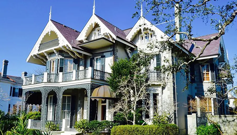 The image features a large, ornate two-story house with multiple gabled roofs, intricate trim details, extensive balconies, and a surrounding garden under a clear blue sky.