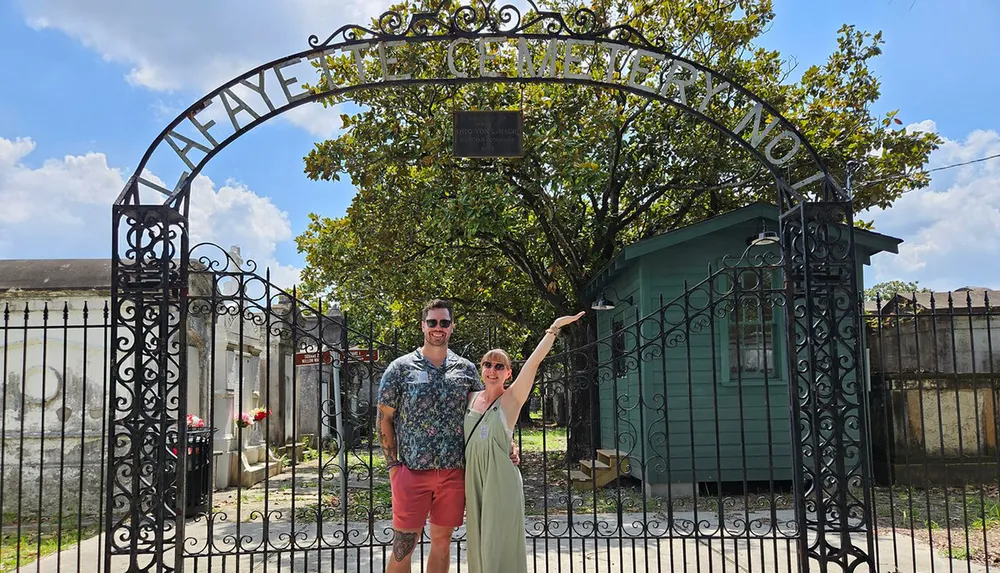 Two people are smiling and posing in front of an ornate cemetery gate under a clear blue sky