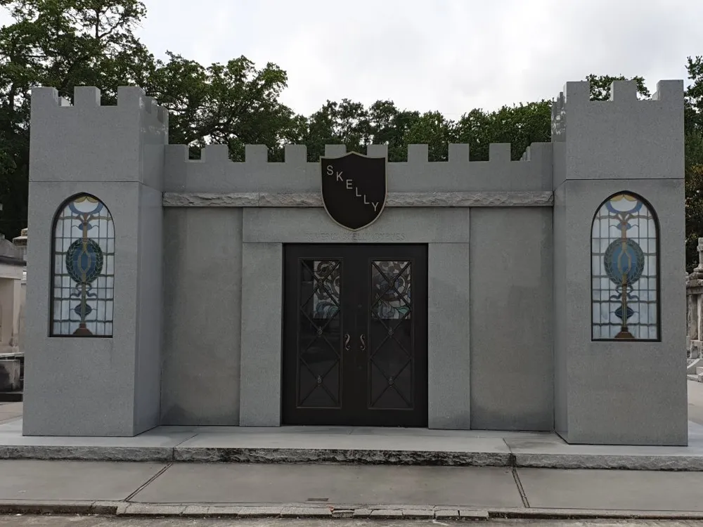 The image shows a modern castle-like mausoleum with gray walls stained-glass windows and a plaque with the name SKELLY