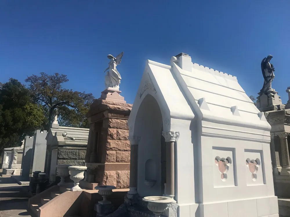 The image shows an arrangement of ornate mausoleums with statues under a clear blue sky in a cemetery
