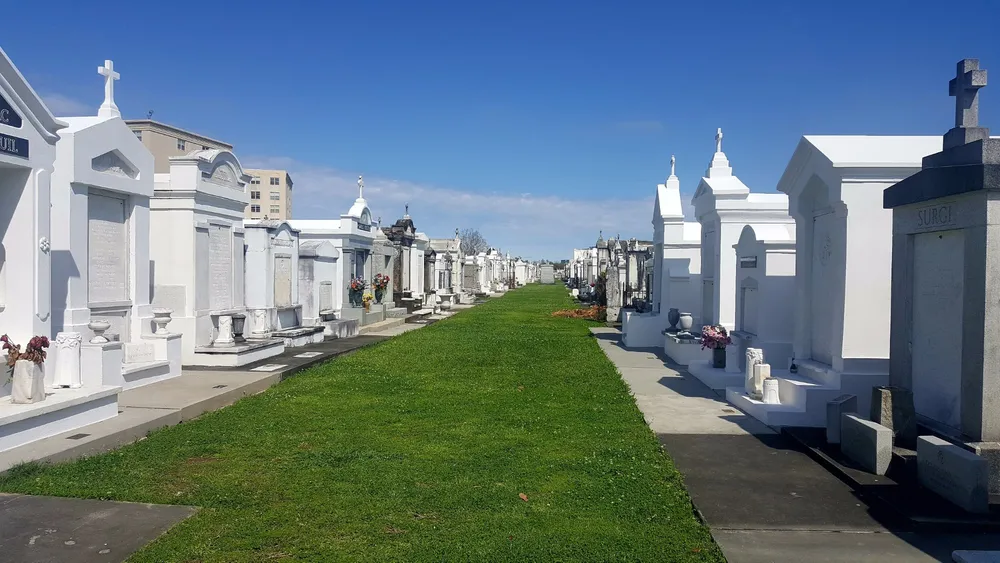 The image shows a pathway flanked by rows of white above-ground tombs and mausoleums under a clear blue sky