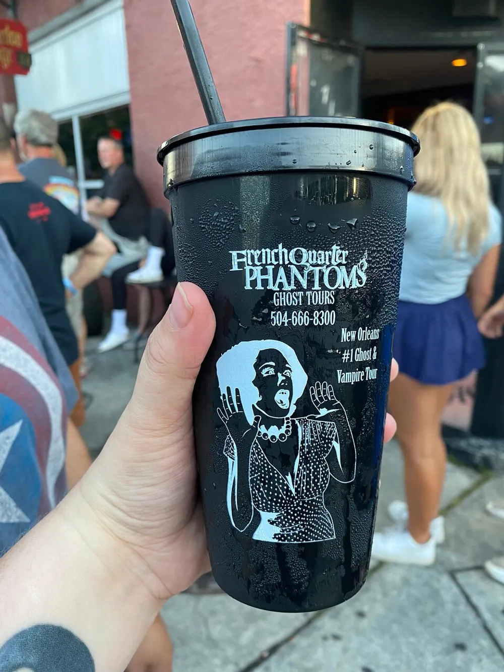 A person is holding a black cup with the French Quarter Phantoms Ghost Tours logo and contact information featuring a graphic of a distressed woman in front of a blurred street scene
