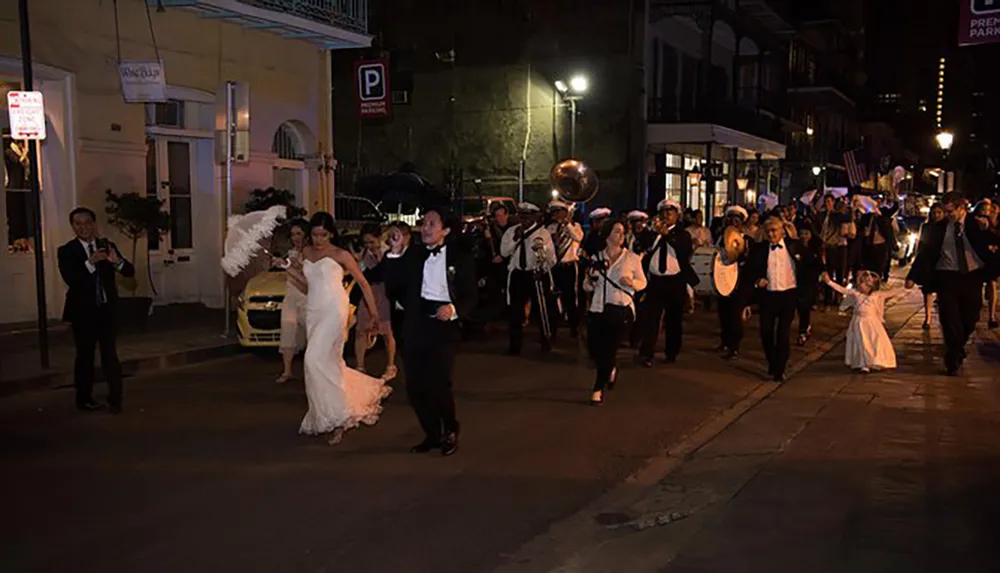 A joyful wedding party led by a bride and groom is followed by a brass band as they partake in a traditional second line parade on an evening street