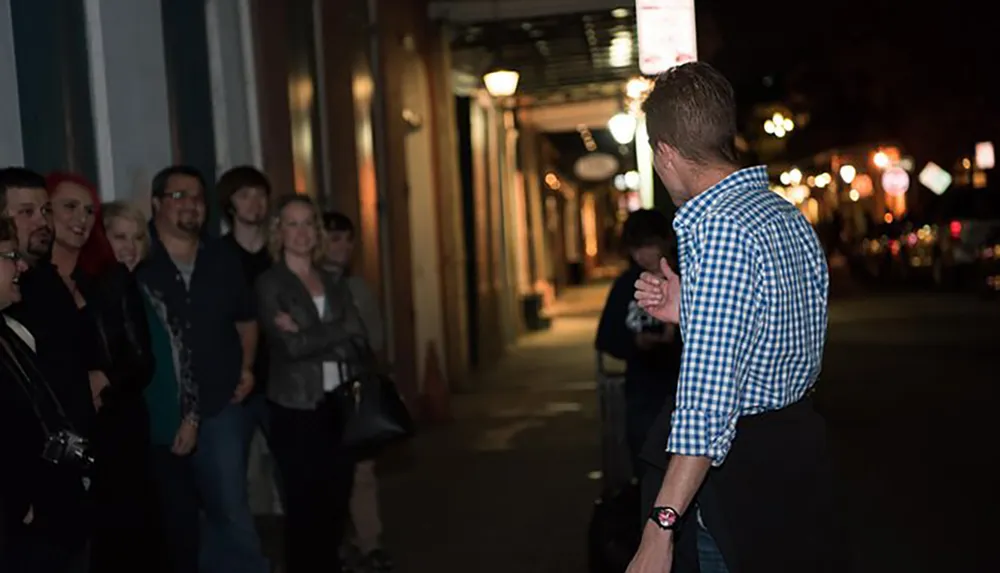 A man in a checkered shirt is leading a group of people down a nightlife-lit street possibly giving a tour