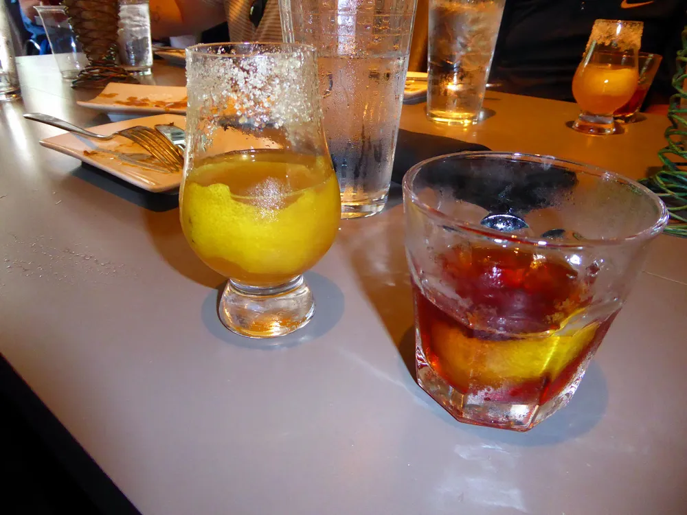 The image shows two different cocktails on a table one with a salty rim and lemon wedge and the other appears to be a whiskey-based drink with both drinks amid a lively dining setting with empty plates and glasses suggesting a group meal