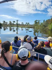 Passengers wearing protective ear gear are on a boat tour, likely an airboat ride, enjoying the tranquil waters and scenic view of a natural landscape, possibly a swamp or wetland area.