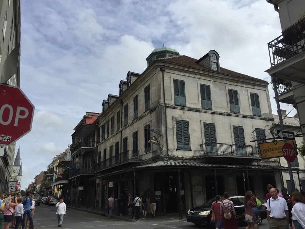 The image shows a bustling street corner in an urban area with pedestrians and classic architecture featuring wrought-iron balconies typical of New Orleans French Quarter