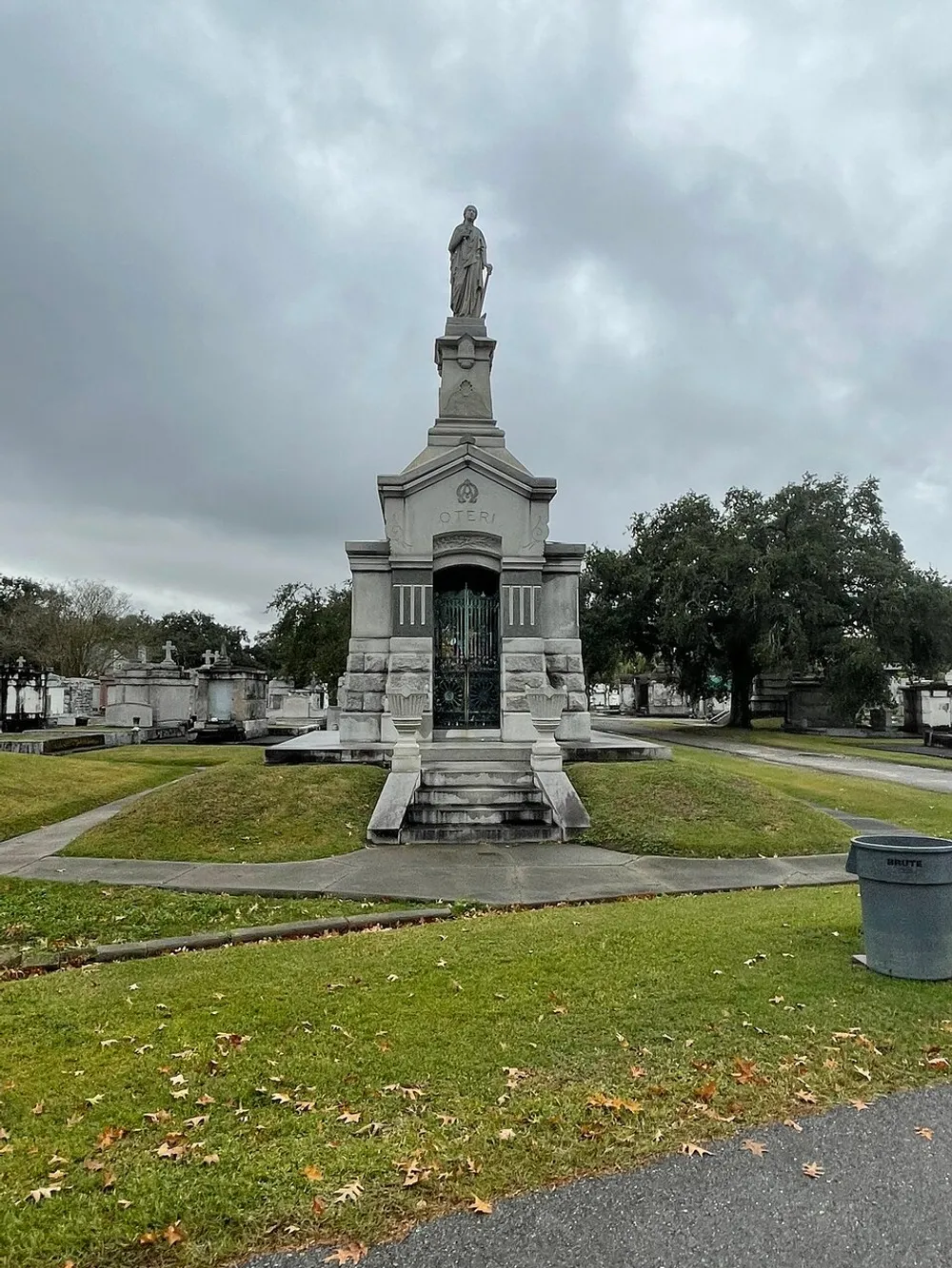 The image depicts an ornate mausoleum with a statue on top under an overcast sky surrounded by a well-maintained lawn dotted with fallen leaves