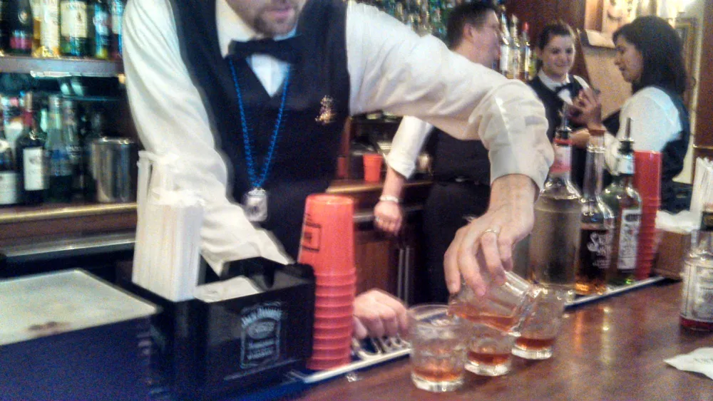 A bartender is preparing drinks at a busy bar with guests in the background