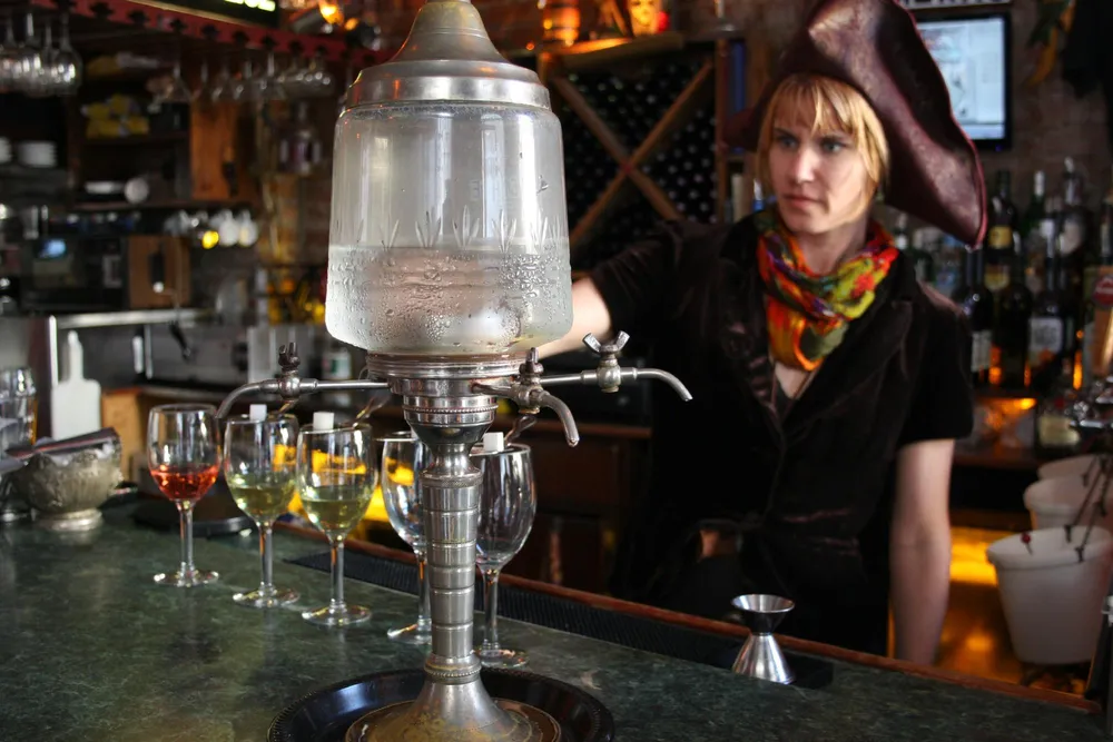 A person dressed in a pirate hat and a scarf is standing behind a bar with a traditional absinthe fountain and glasses filled with varying amounts of absinthe