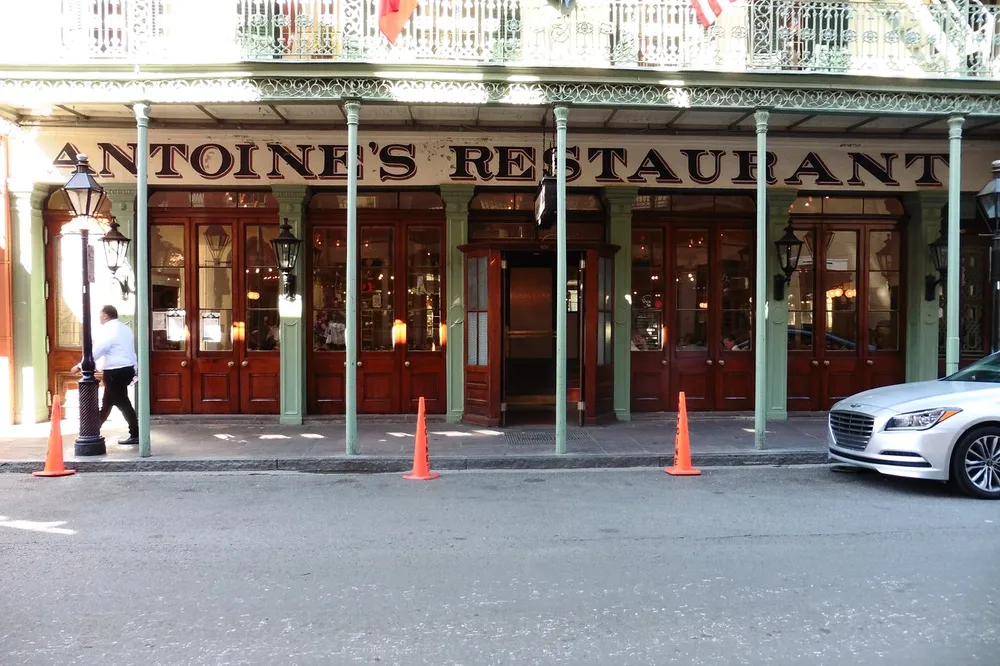 The image shows the exterior of Antoines Restaurant with its classic wooden doors and iron balcony with a person walking by and a car parked on the street