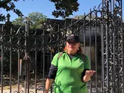 A person is standing in front of an ornate metal gate that appears to be the entrance to a cemetery or crypt, gesturing with an open hand.