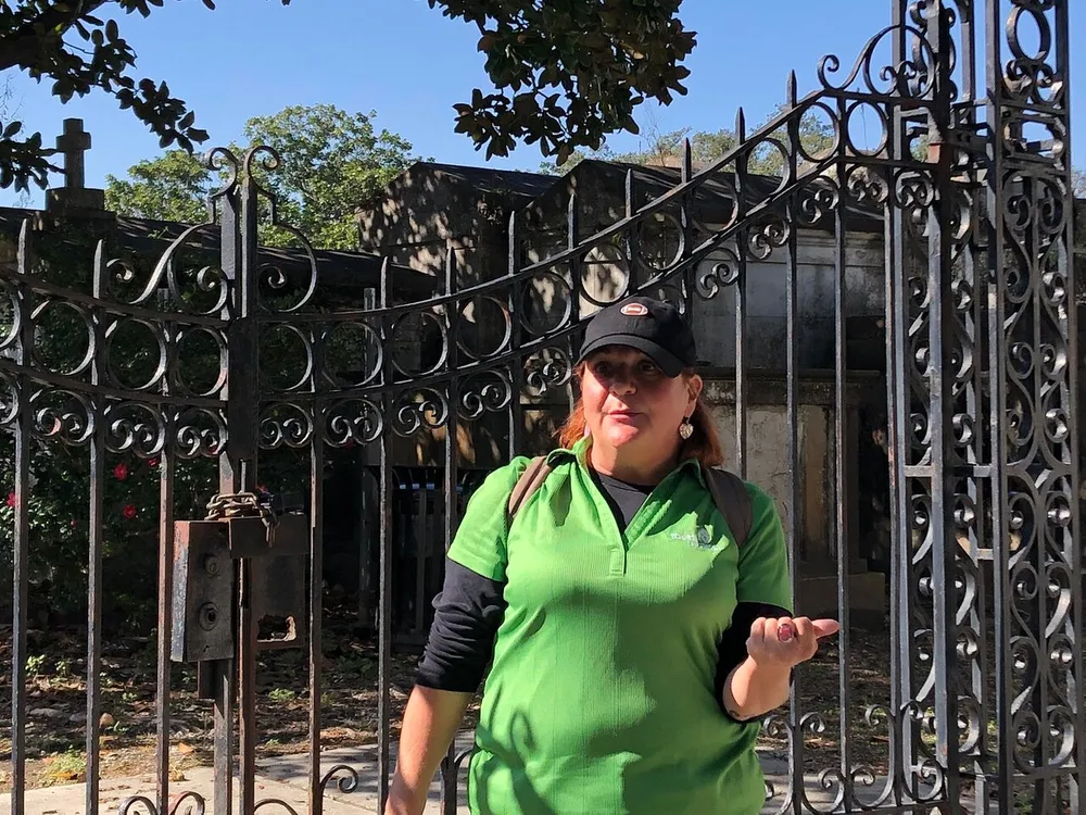 A person is standing in front of an ornate metal gate that appears to be the entrance to a cemetery or crypt gesturing with an open hand