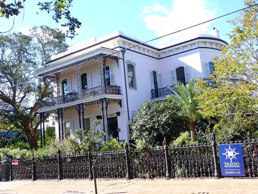 The image shows a two-story white house with ornate black ironwork on the balconies surrounded by lush greenery and a wrought-iron fence displaying the character of traditional southern architecture