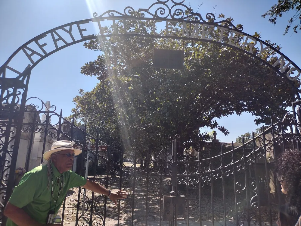 A person in a green shirt and hat is gesturing while standing by an ornate gate with the words LAFAYETTE CEMETERY on it in the presence of another individual under a sunny sky