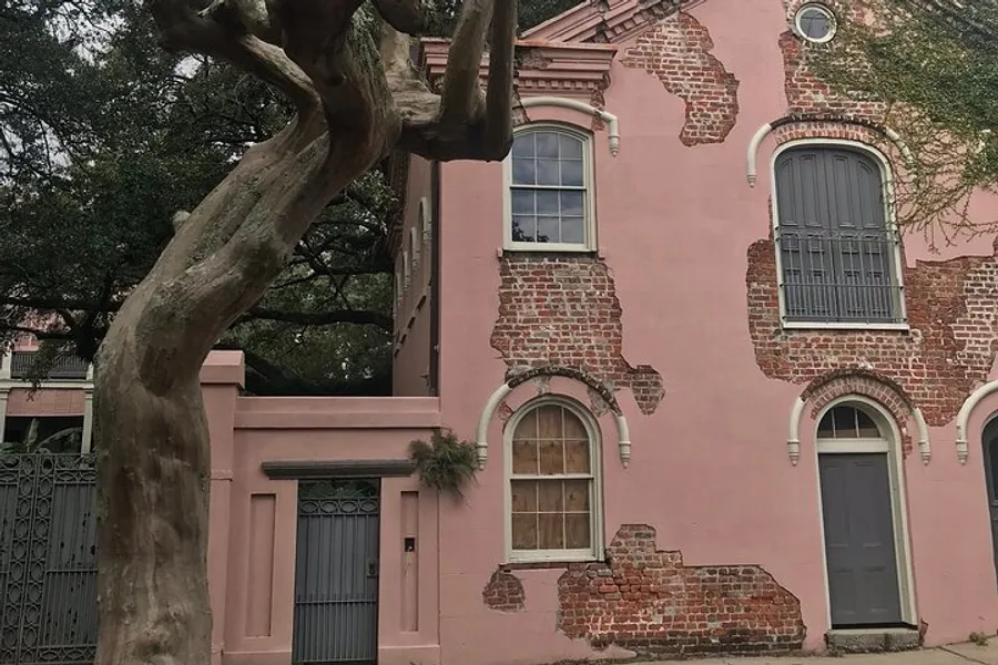 The image shows an aged pink brick building with a large tree in front, featuring elements of peeling plaster and exposed bricks, contributing to a sense of historic charm.