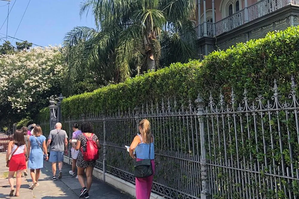 Tourists are walking past a wrought iron fence surrounding a lush garden in front of an ornate building on a sunny day