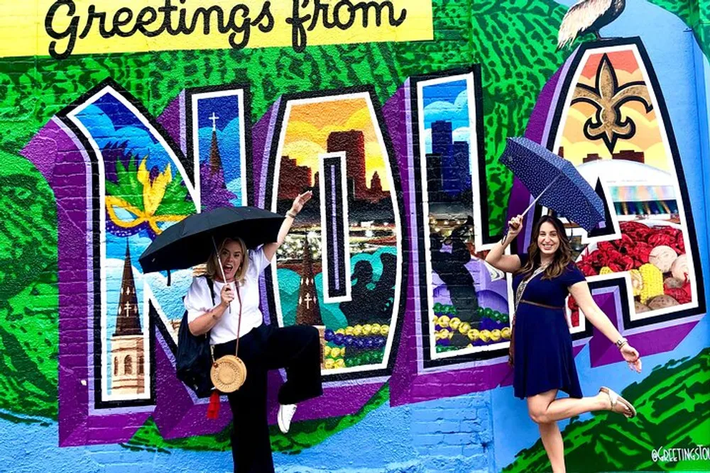 Two people are posing joyfully in front of a colorful mural that says Greetings from NOLA with one holding an umbrella and the other extending their leg in a playful manner