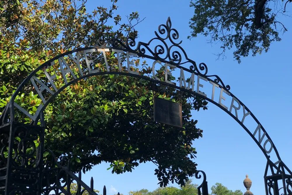 This is an ornate wrought-iron gate with the word CEMETERY on its arch set against a backdrop of a clear blue sky and surrounded by lush green foliage