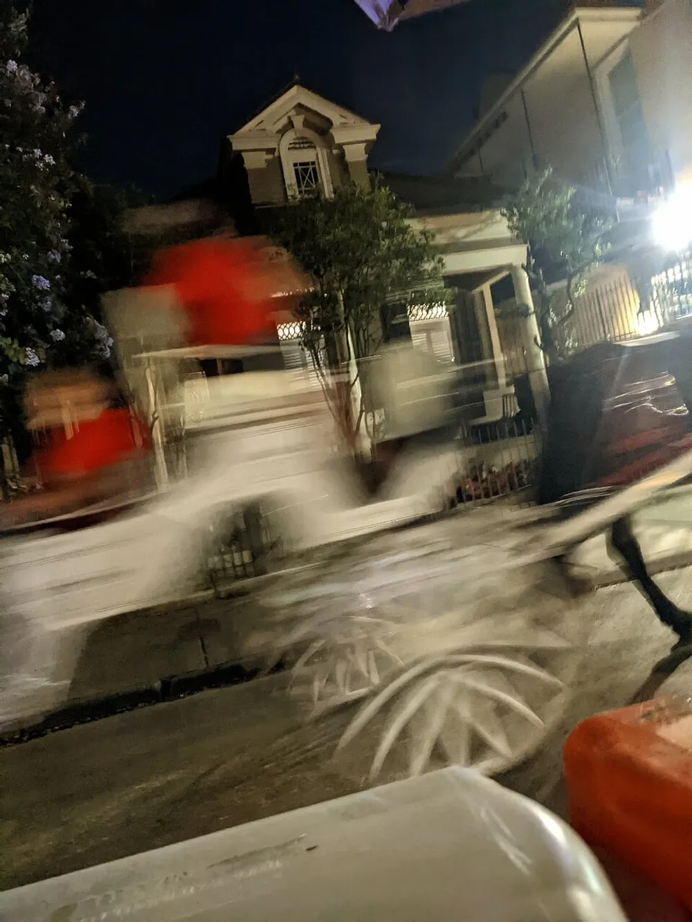 The image shows a blurred scene on a street at night with a house in the background creating a sense of motion likely from a moving vehicle or camera shake