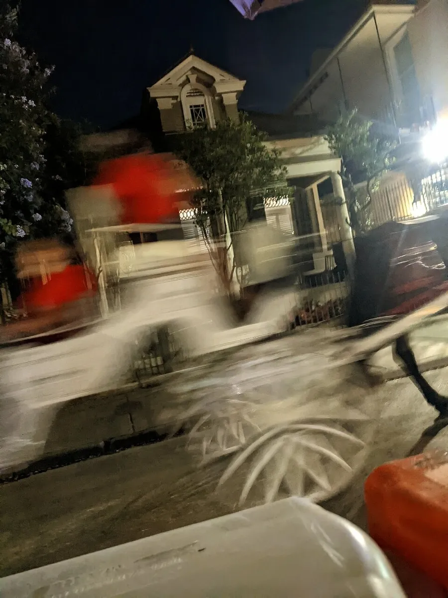 The image shows a blurred scene on a street at night with a house in the background, creating a sense of motion, likely from a moving vehicle or camera shake.