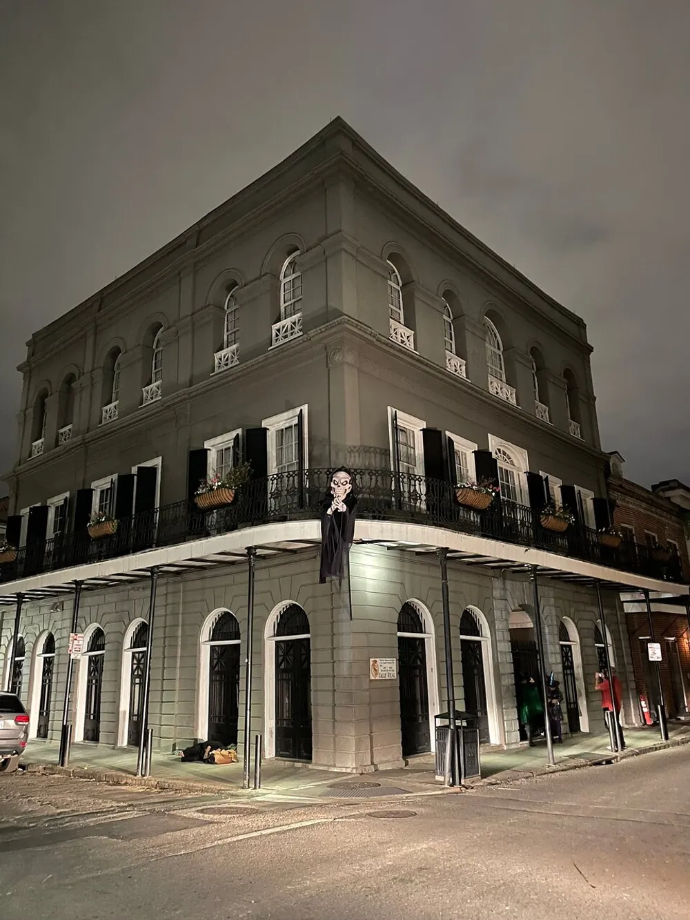 The image shows a two-story corner building at night with a figure resembling a skeleton hanging from a balcony creating a haunting and eerie atmosphere