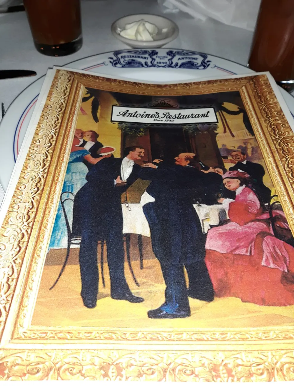 The image shows a menu cover from Antoines Restaurant featuring a colorful vintage illustration of elegantly dressed people dining with a plate bordered with the restaurants name in front of it and a glass of iced tea and a small bowl of butter behind it