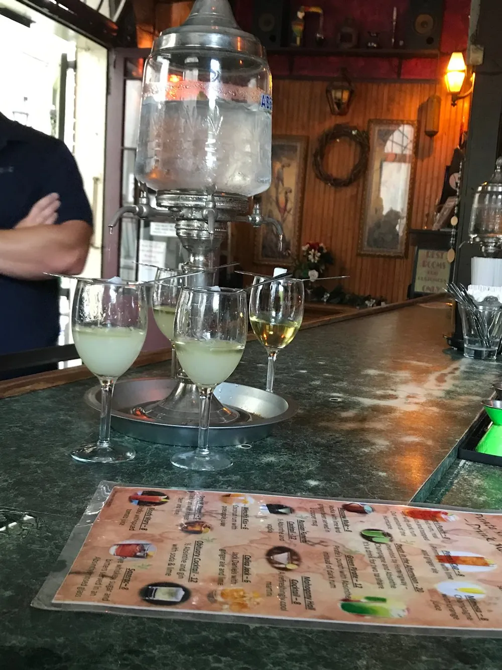 The image shows a bar scene with a drinks menu in the foreground glasses of beverages on the counter and a person standing behind the bar partially cropped out