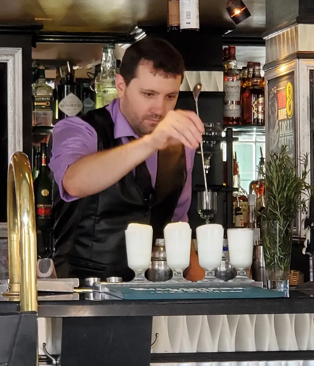 A bartender is intently garnishing or preparing multiple frothy drinks behind a bar with various bottles and bar equipment in the background