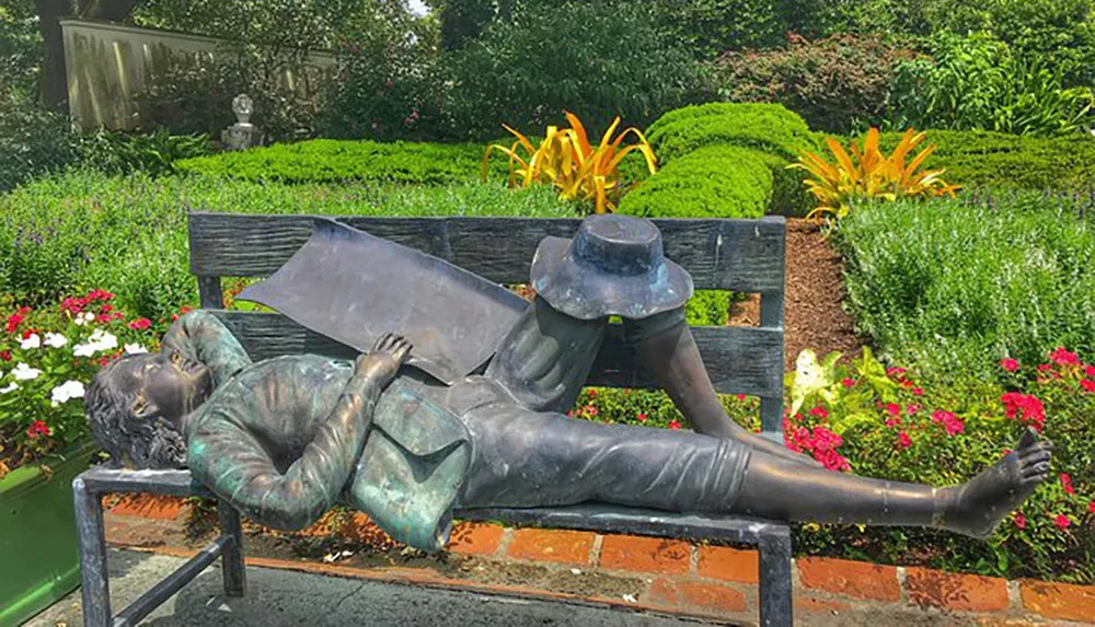 The image features a bronze statue of a person lying on their back on a park bench holding a book above their face set against a lush garden backdrop