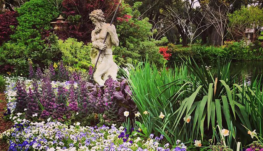 A statue stands amidst a colorful garden with a variety of flowers and shrubs by a pond
