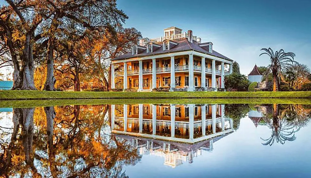 The image shows a stately historic mansion with a large porch and tall columns set against a backdrop of mature trees and reflected perfectly in a still body of water in the foreground