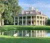 A grand plantation-style house with a two-story columned facade overlooks a serene pond with two swans in the foreground amidst lush greenery