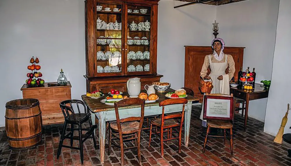 The image shows a rustic kitchen scene with wooden furniture a variety of foods displayed on the table and a mannequin dressed in period attire suggesting an historical or museum setting