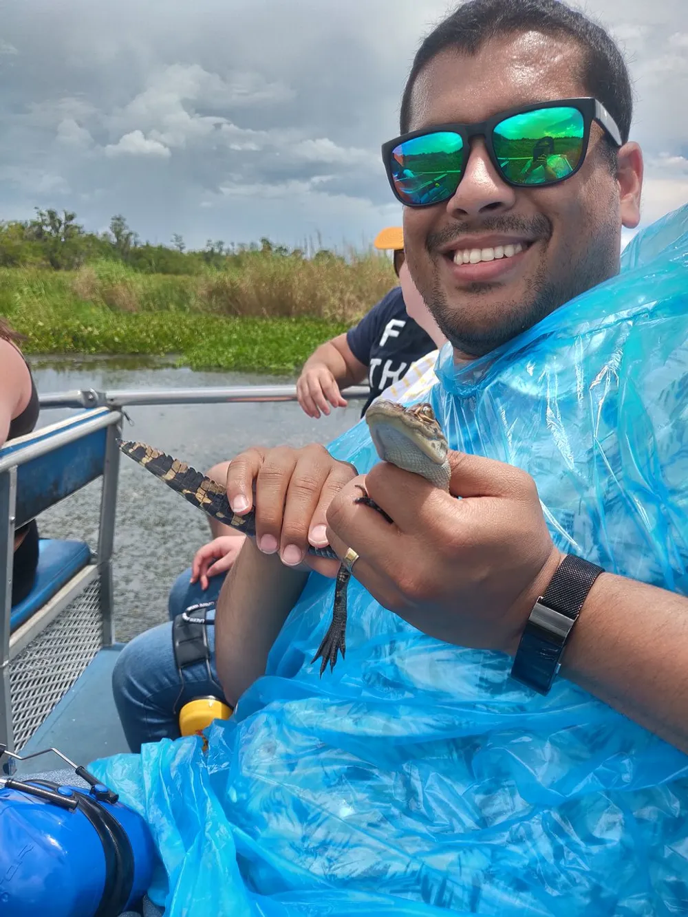 A person wearing a blue poncho and sunglasses is smiling while holding a baby alligator on a boat