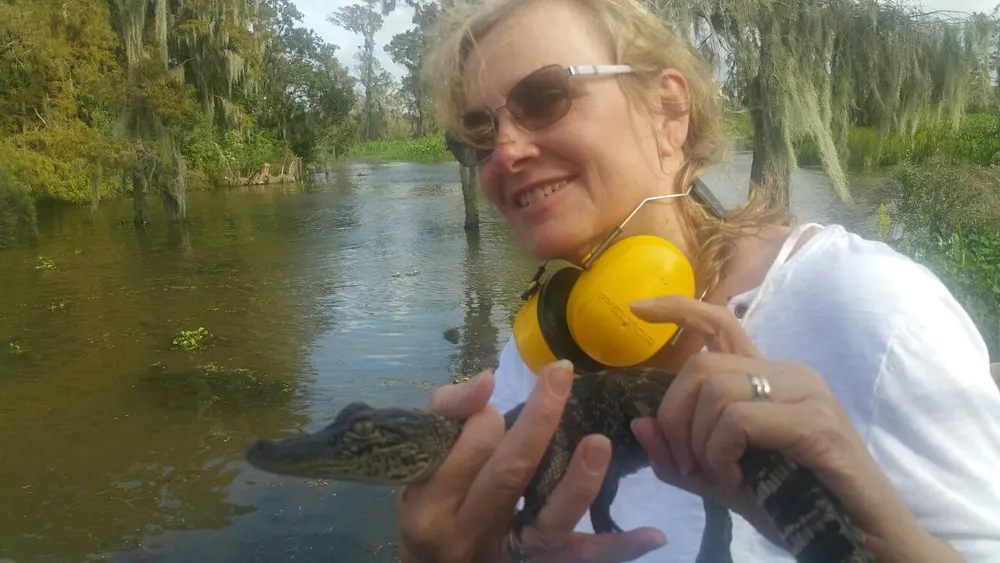 A person is holding a young alligator with a smile wearing headphones around their neck with a marshy waterway in the background