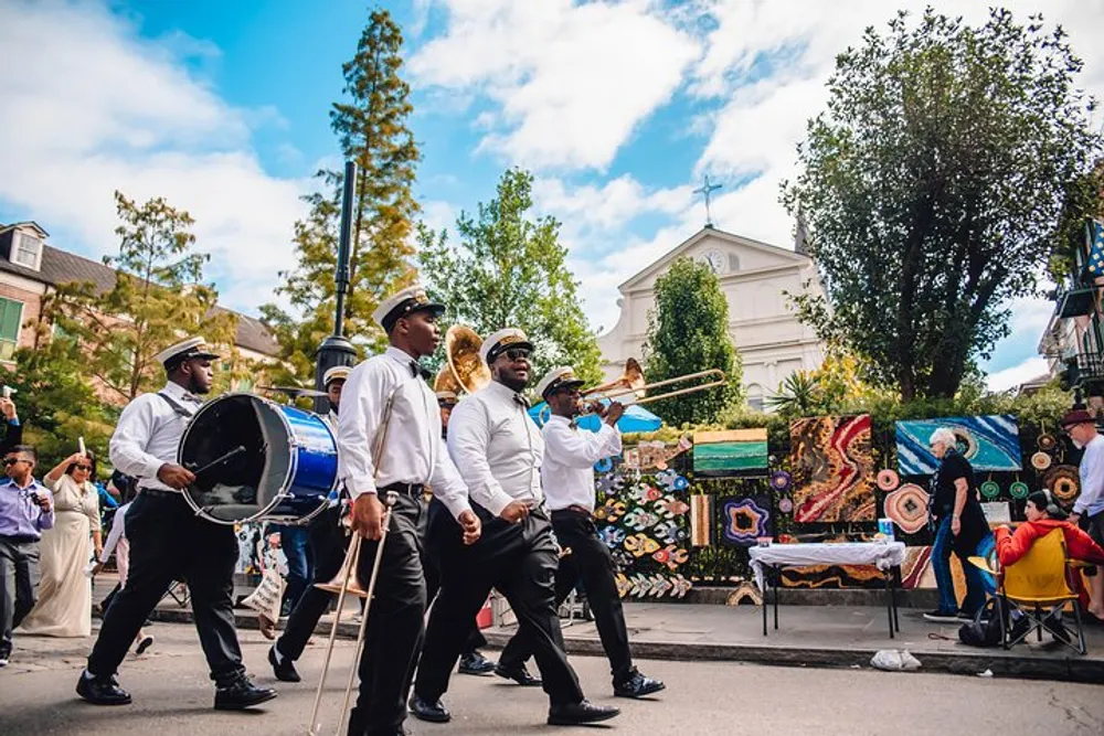 A brass band dressed in white shirts and black pants with some members wearing hats is playing their instruments while participating in a street parade with onlookers and colorful decorations around them