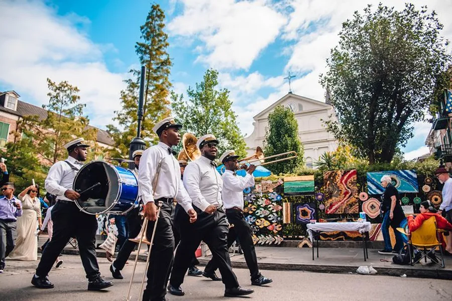 A brass band, dressed in white shirts and black pants with some members wearing hats, is playing their instruments while participating in a street parade with onlookers and colorful decorations around them.