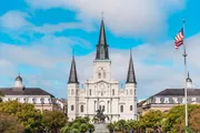 The image features the iconic Saint Louis Cathedral with its white facade and three spires, overlooking Jackson Square in New Orleans, under a blue sky, with a statue in the foreground and the American flag to the right.