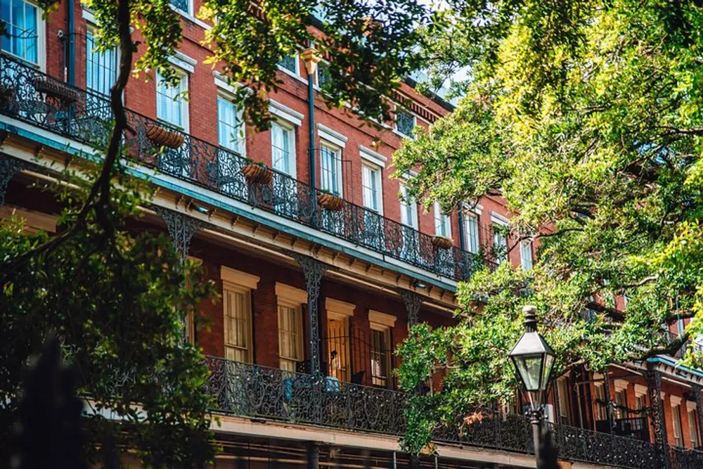 The image shows a street view of a row of traditional red brick buildings with ornate black ironwork on the balconies partially obscured by leafy green trees evoking a sense of historic urban charm