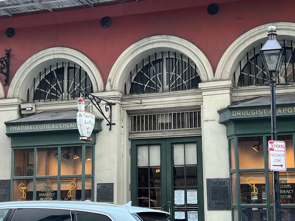 The image shows the exterior of the New Orleans Pharmacy Museum with its distinctive signage and a mortar and pestle icon against a backdrop of red and white architecture and a No Parking sign visible by the roadside