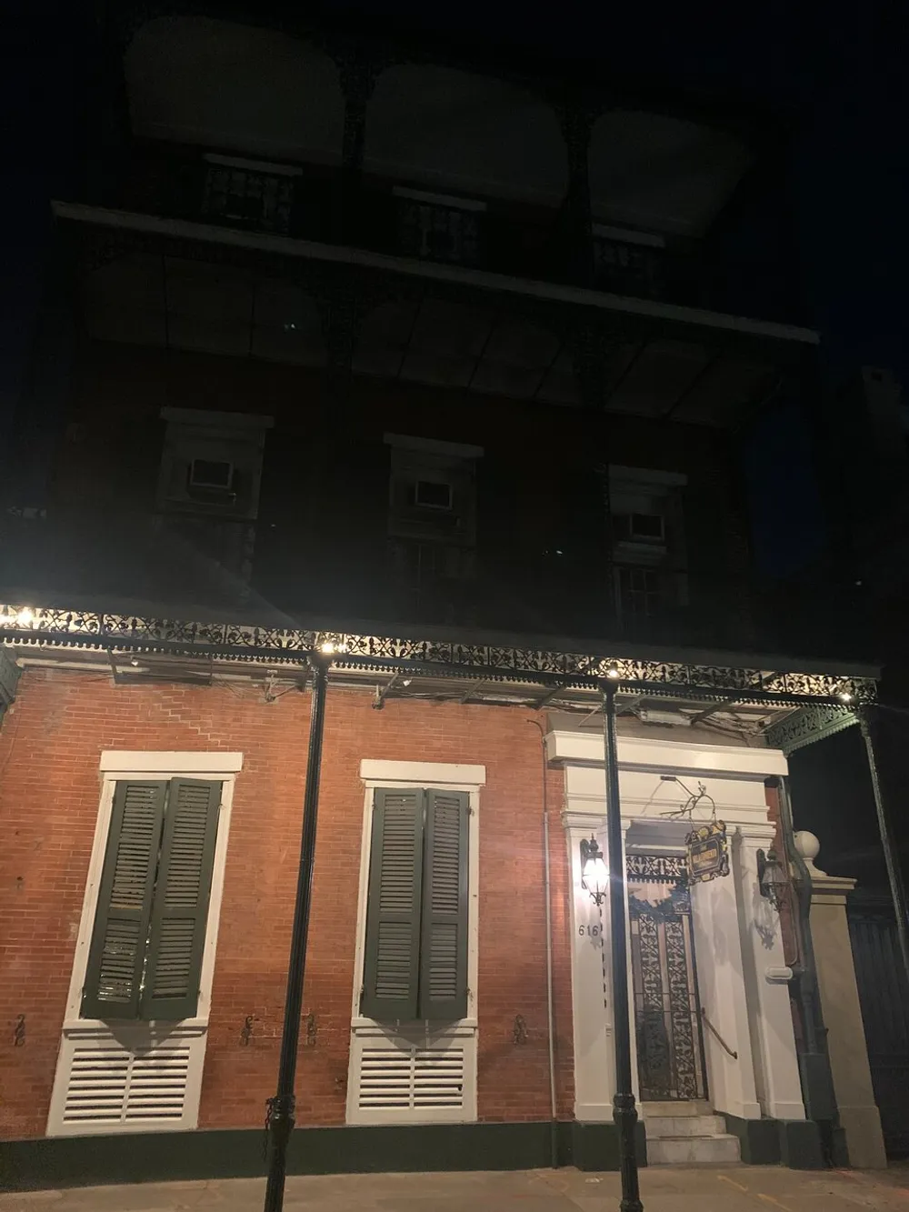 The image shows a nighttime view of a two-story red brick building with white shutters ornate wrought iron balcony details and a street lamp in the foreground