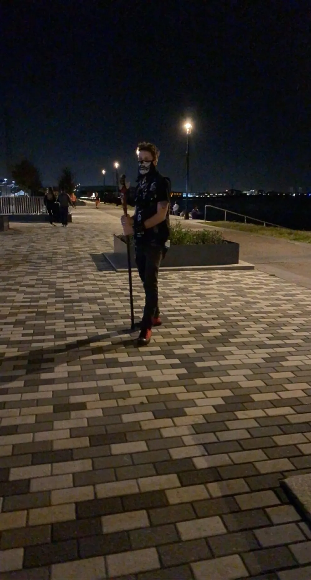 A person wearing a mask is standing on a paved walkway at night holding a walking stick with streetlights and people in the distant background