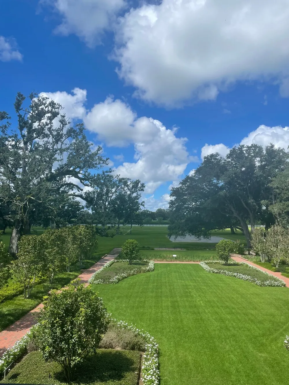 The image shows a well-maintained garden with manicured lawns bordered by neatly trimmed hedges and trees with a brick pathway leading towards a water body under a bright blue sky scattered with clouds