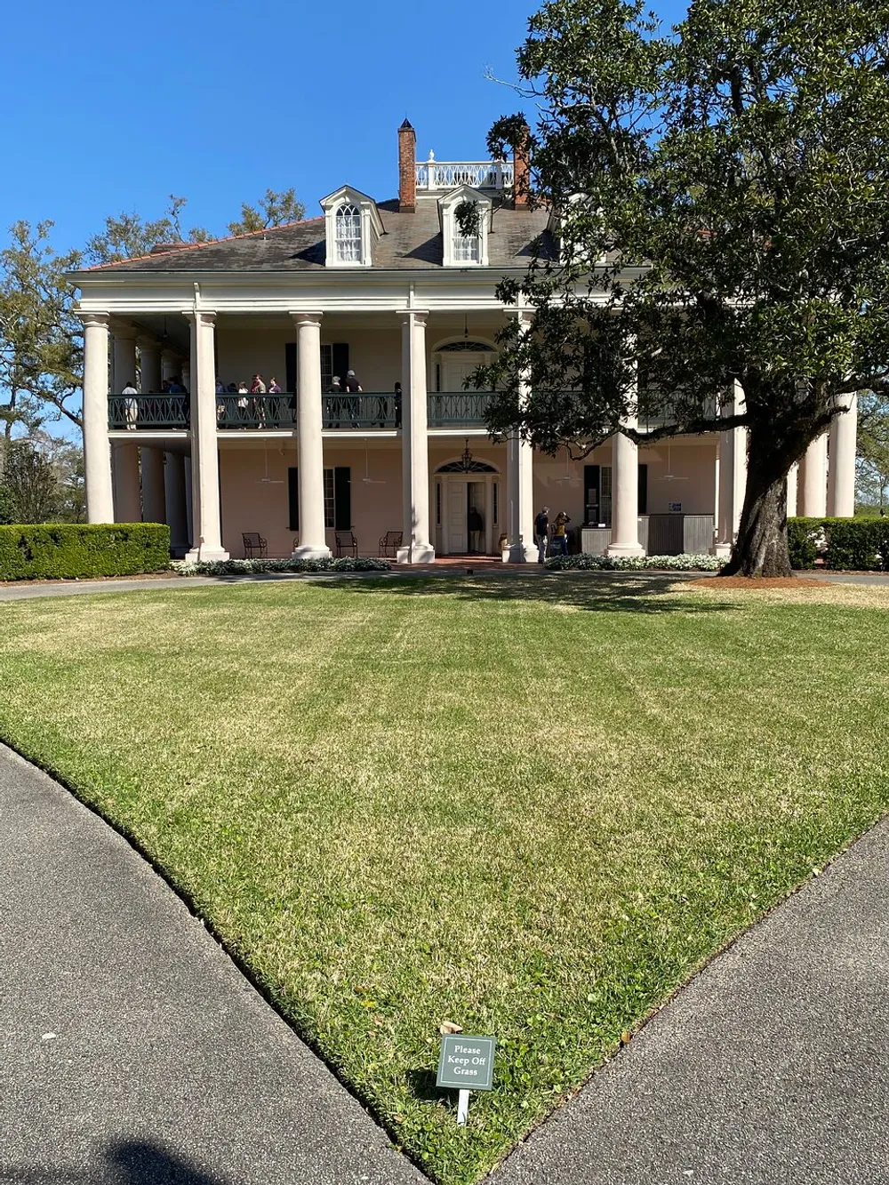 The image displays a grand two-story house with a large porch and columns surrounded by a well-manicured lawn with a sign reading Please Keep Off Grass and a few visitors visible on the upper balcony