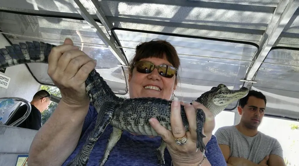 A smiling woman wearing sunglasses is holding a small alligator on a covered boat with a man visible in the background
