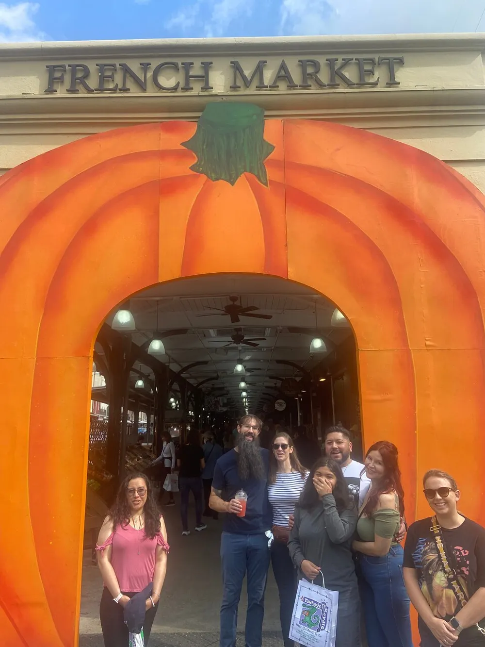 A group of individuals poses for a photo in front of the entrance to the French Market which is adorned with a large decorative pumpkin facade