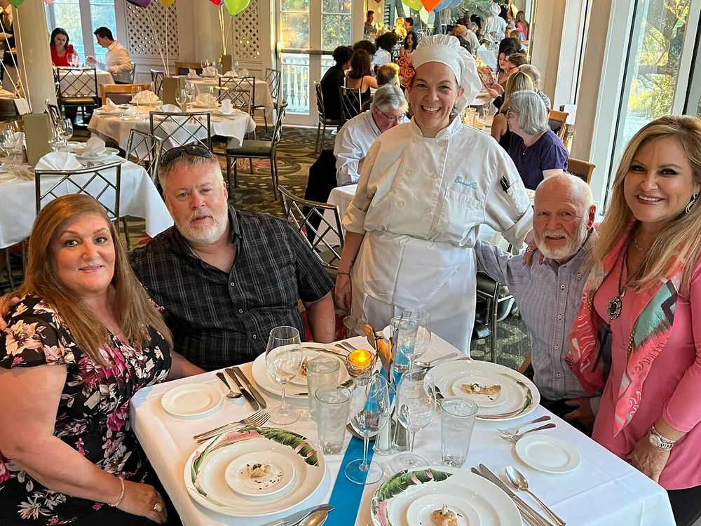 Four diners are posing with a smiling chef at a well-set table in a restaurant bustling with activity