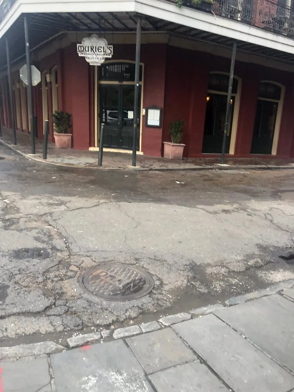 The image shows a red-colored building with an establishment named Muriels under a covered sidewalk with signs of wear on the street infrastructure including a pothole and a manhole cover