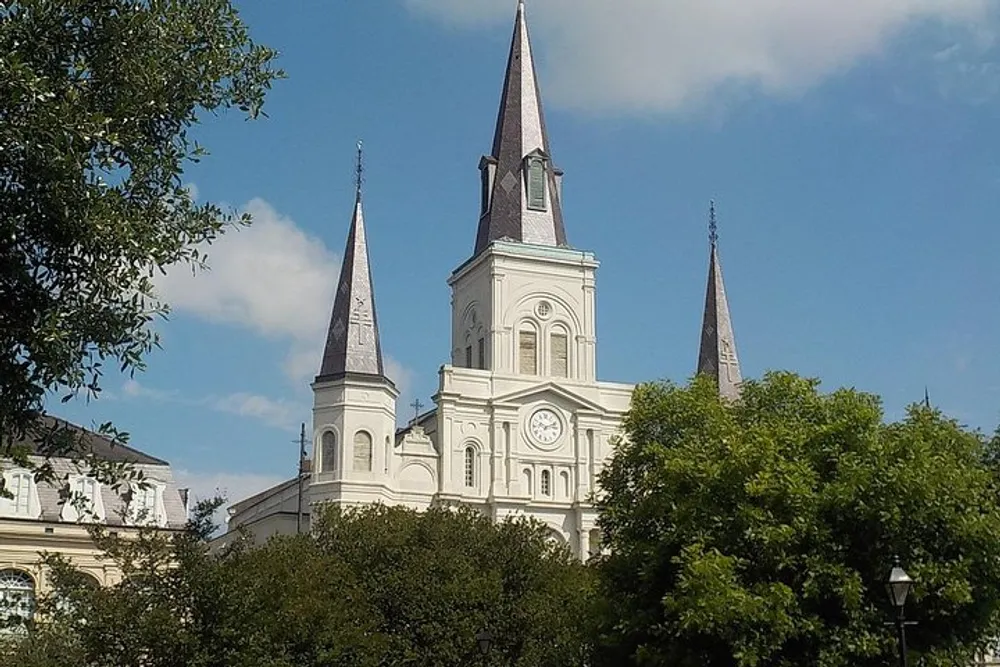 The image shows the iconic triple steeples of a white cathedral rising above the treeline against a blue sky with some white clouds
